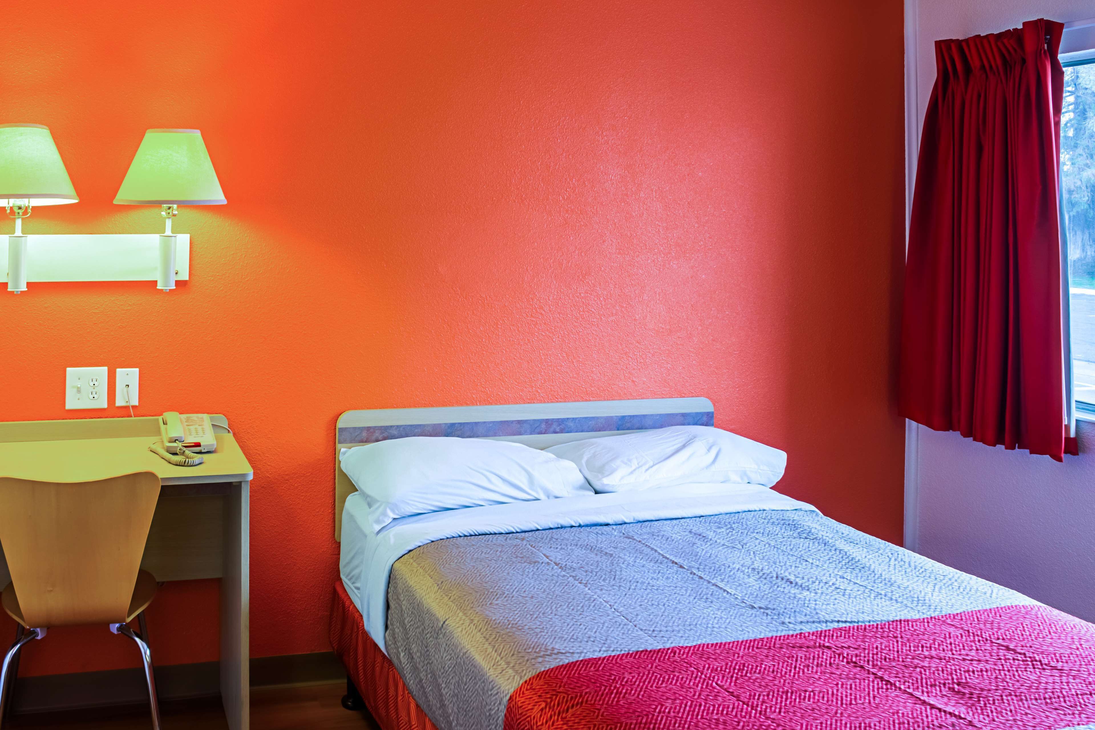 Una habitación con cama, escritorio, dos lámparas y ventana con cortinas rojas.