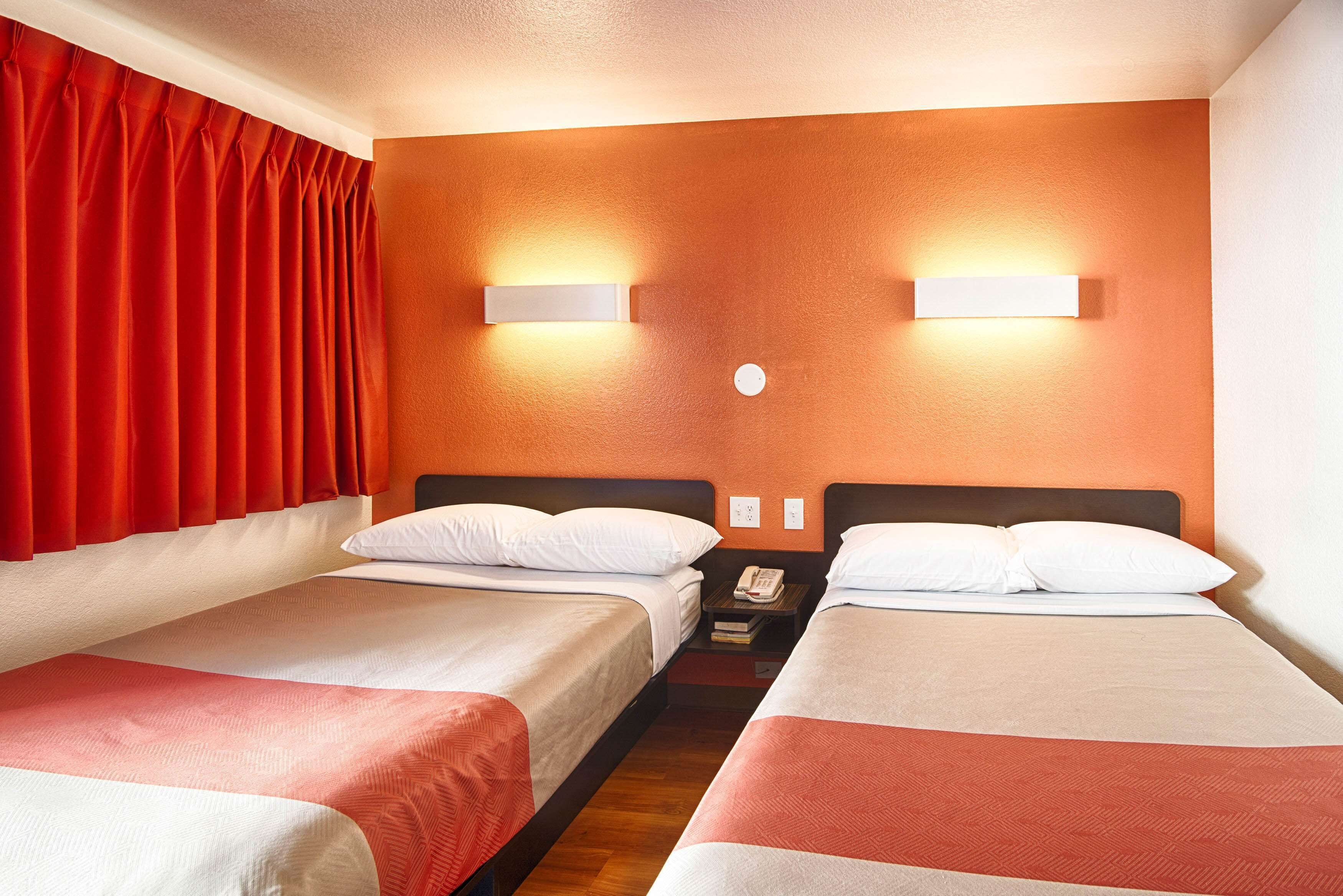A hotel room with two single beds, a nightstand with a phone, and a wall-mounted light fixture.