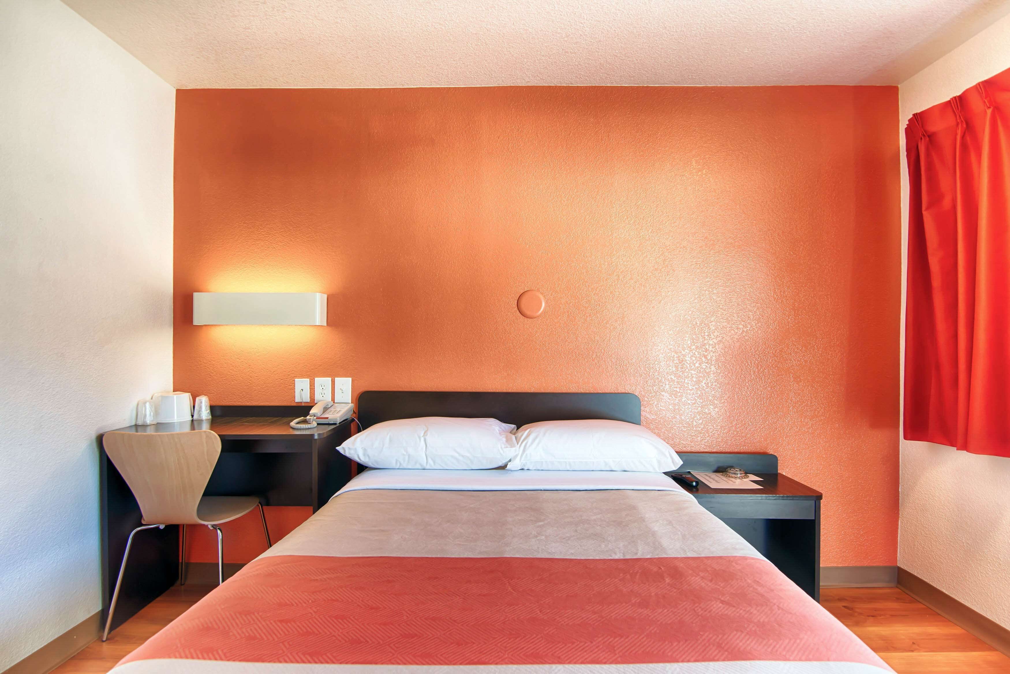 Une chambre d’hôtel équipée d’un lit, d’un bureau, d’une chaise, d’une table de chevet et d’une lampe fixée au mur.