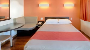 Quarto de hotel com cama, escrivaninha, cadeira e duas luminárias fixadas na parede.