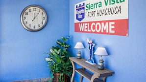 Un reloj, un cartel de bienvenida, una planta en maceta y una mesa con dos lámparas.