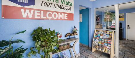 A hotel reception area with a sign for Knights Inn Sierra Vista Fort Huachuca, a welcome sign, a bulletin board with travel brochures, and a plant on a wooden stand.