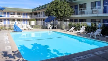 A swimming pool with lounge chairs and umbrellas, surrounded by a fence, with a building in the background.