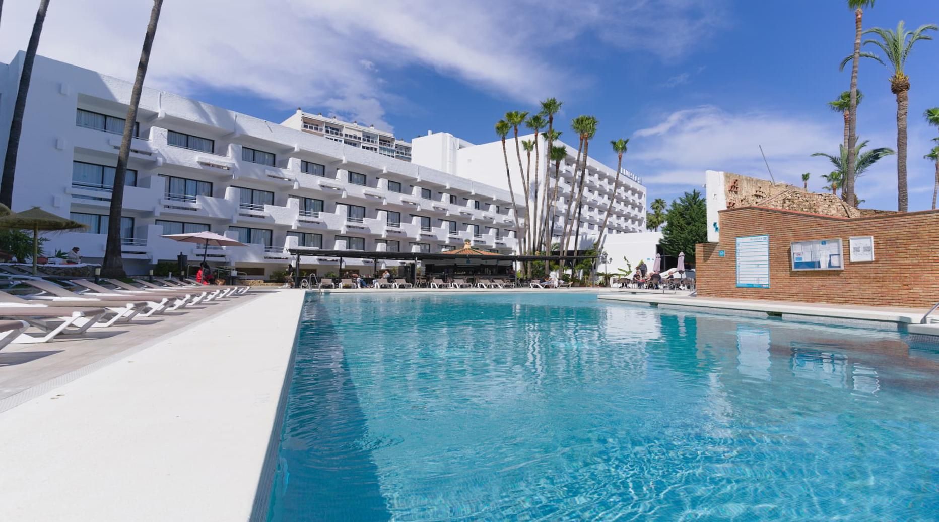 A hotel with a pool, palm trees, and a clear sky.