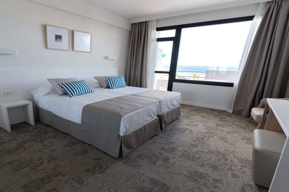 Une chambre d’hôtel avec un grand lit, une table de chevet, un petit bureau et une vue sur la mer par la fenêtre.