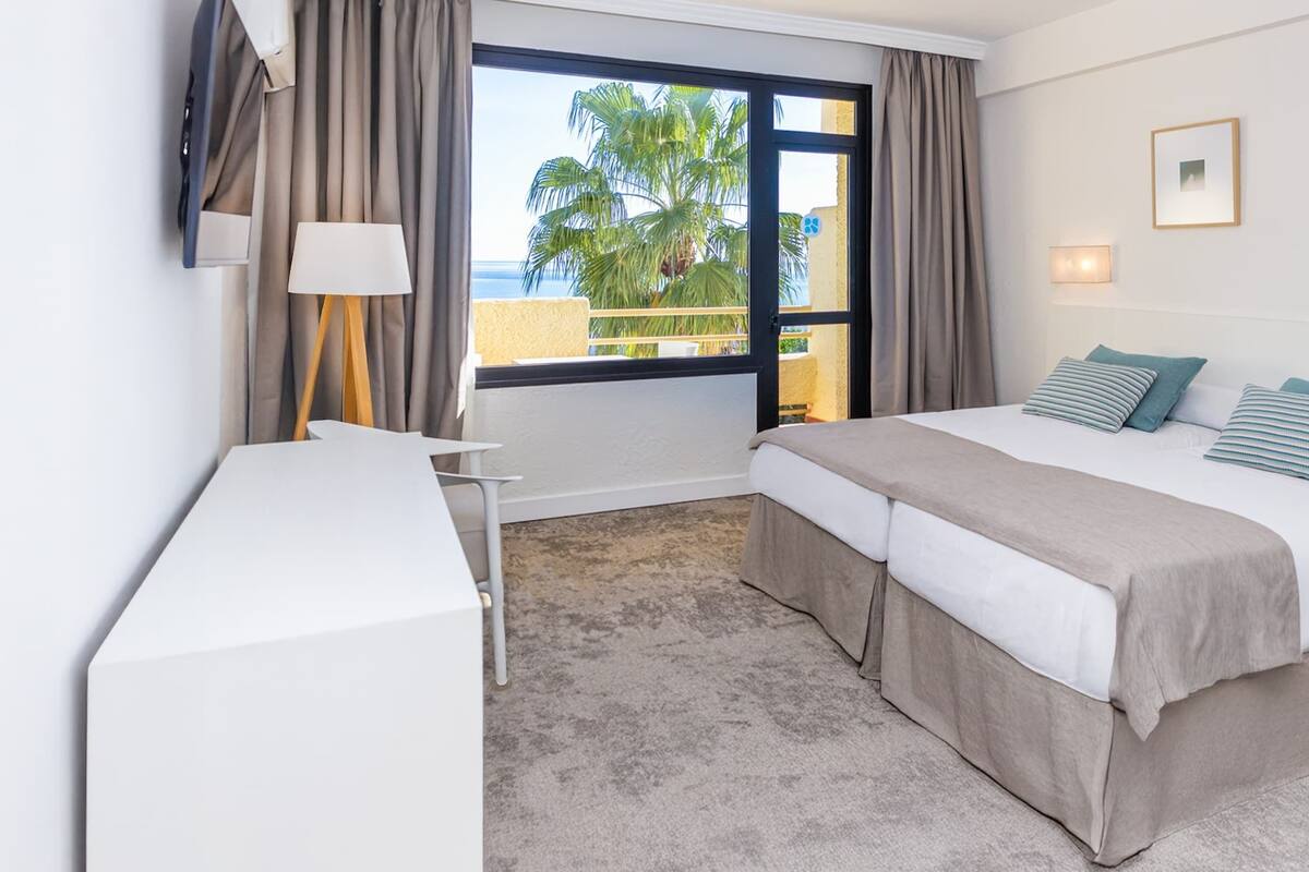 Une chambre d’hôtel avec un lit, un bureau et une vue sur les palmiers et la mer.