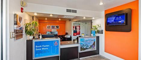 A hotel reception area with a modern design, featuring a reception desk, a digital screen displaying information, and a sign promoting a mobile app for booking.