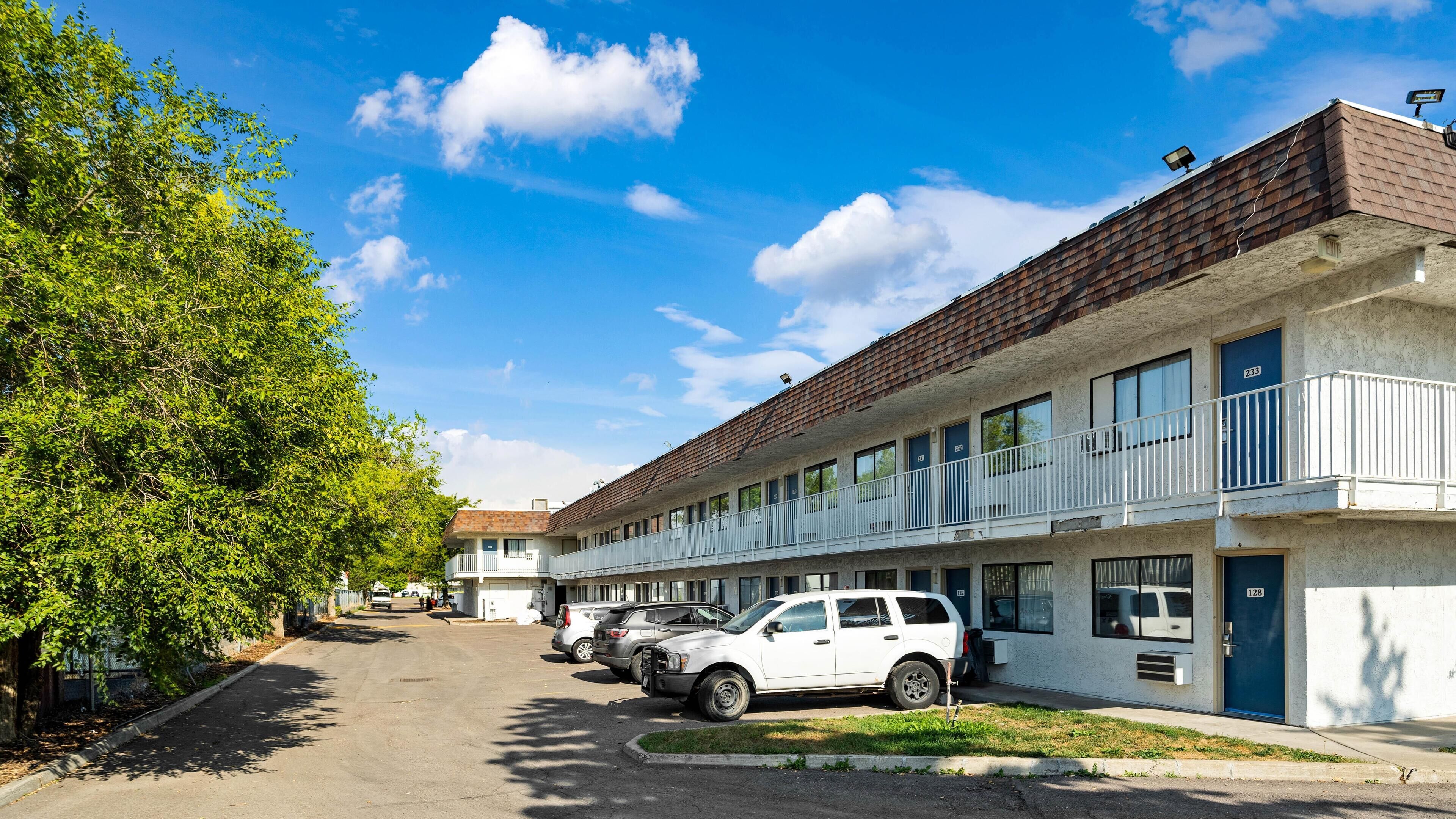 Una fila di edifici motel a due piani con balconi, auto parcheggiate e una strada alberata.