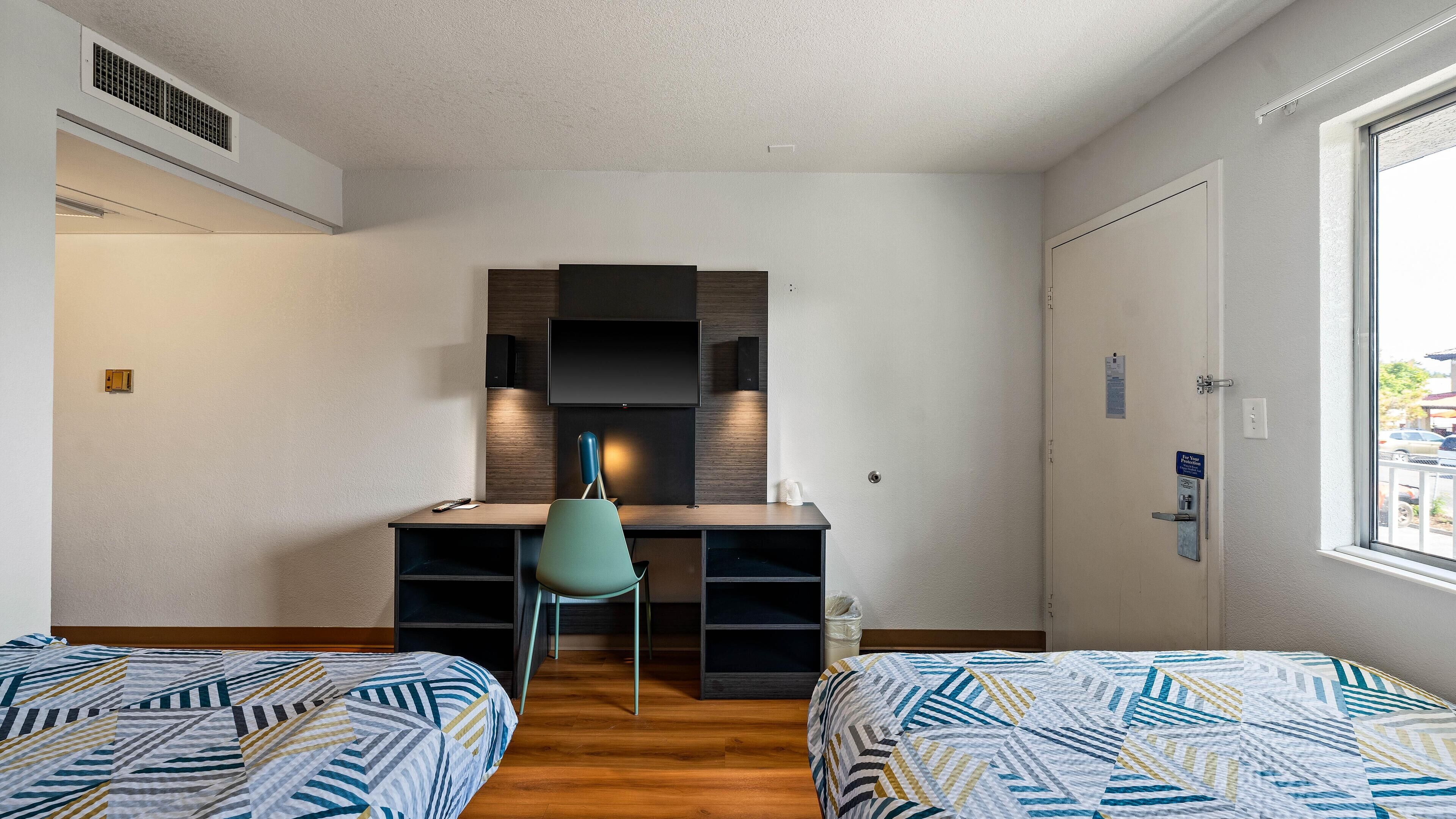 A bedroom with two beds, a desk with a chair, a TV, and a window.
