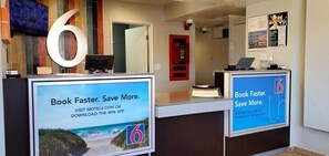 A reception desk with a large number '6' sign, a computer, and promotional posters for booking through the motel's website or app. - Motel 6 Palmdale, CA (Palmdale)