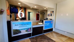 A reception desk with a large number '6' sign, two digital screens displaying promotional messages, and a tiled floor. - Motel 6 Palmdale, CA (Palmdale)