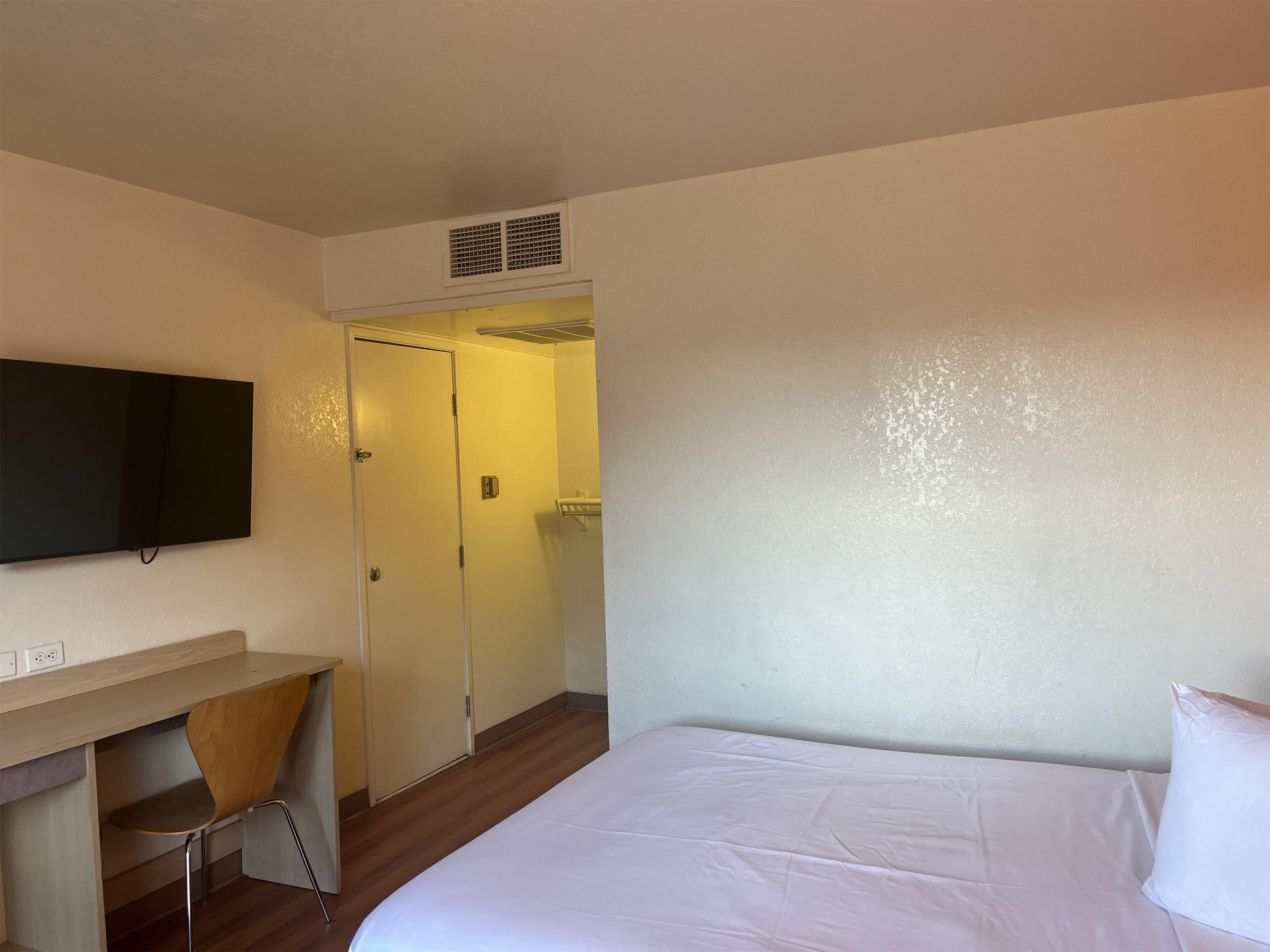 A hotel room with a bed, a desk with a chair, a TV mounted on the wall, and a door leading to a bathroom.
