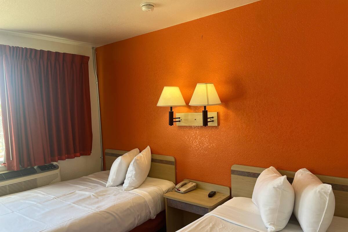 Habitación de hotel con dos camas, una mesita de noche con teléfono y control remoto, y una lámpara de pared.