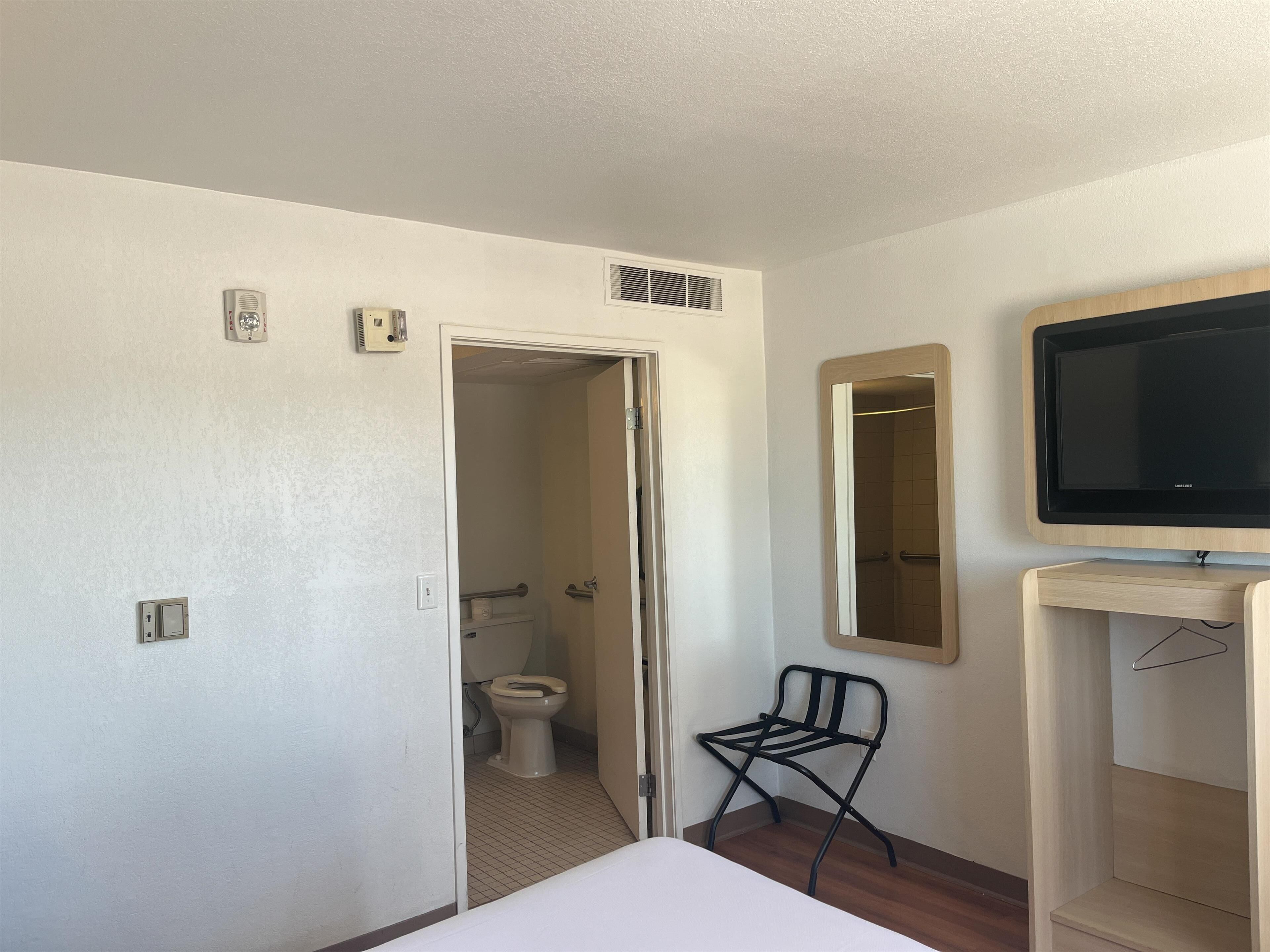 A hotel room with a bed, a television on a wooden stand, a mirror, a chair, and a bathroom visible through an open door.