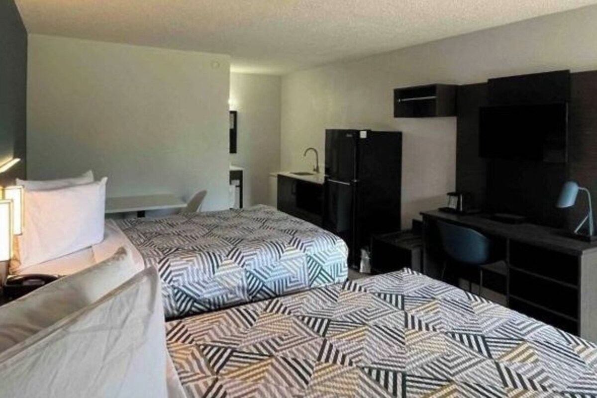 Quarto de hotel com duas camas, uma escrivaninha, uma cadeira, uma televisão e uma cozinha compacta.
