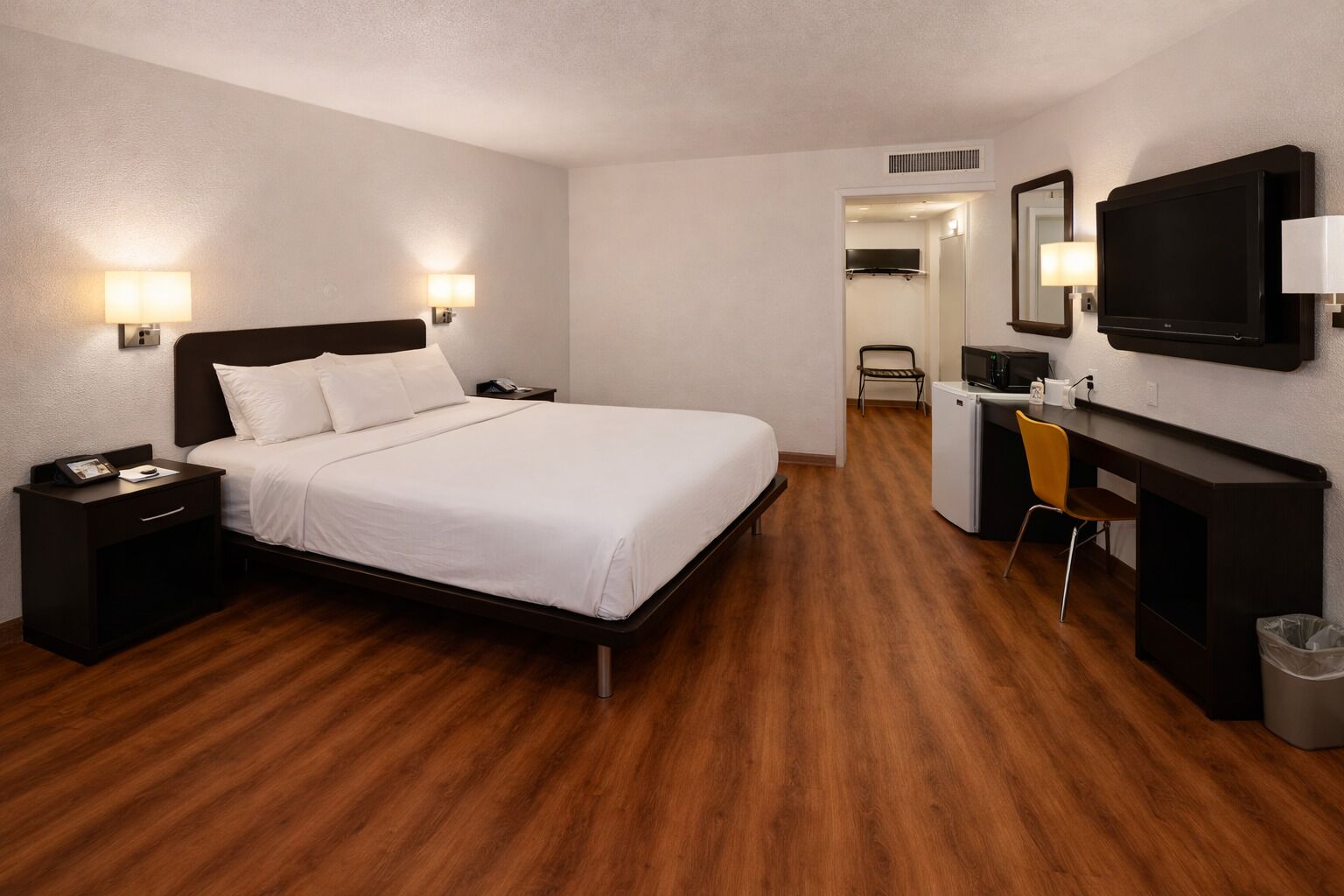 A hotel room with a bed, bedside tables, a desk with a chair, a TV, and a trash bin.