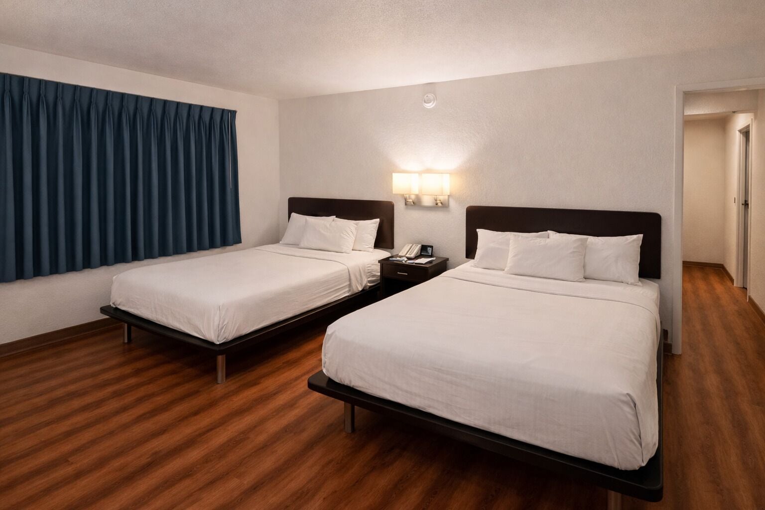 Una habitació d'hotel amb dos llits individuals, terra de fusta, una cortina blava i una taula de nit amb una làmpada.