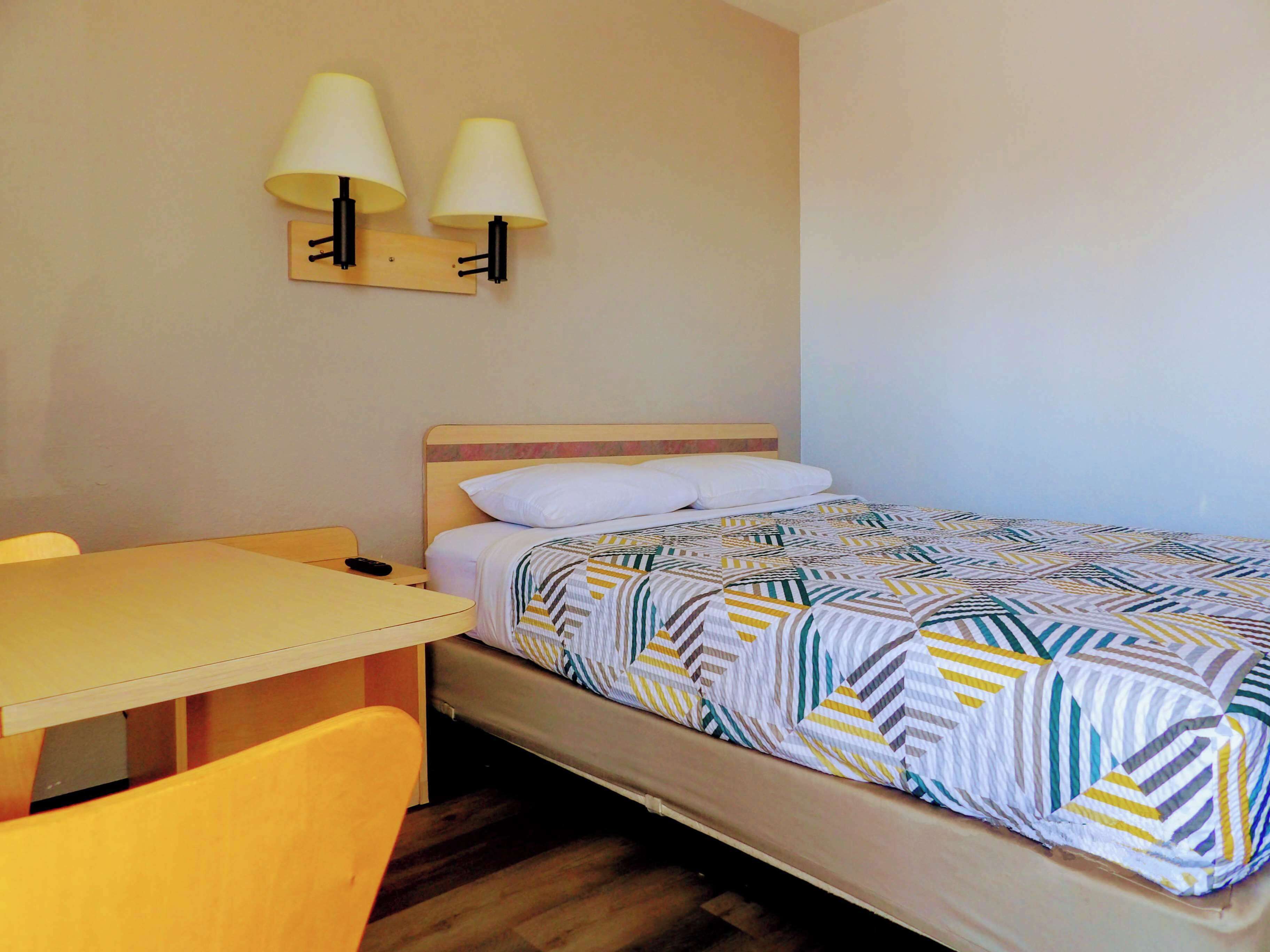 Una cama individual con una colcha estampada, una mesita de noche, una silla y dos lámparas fijadas a la pared.