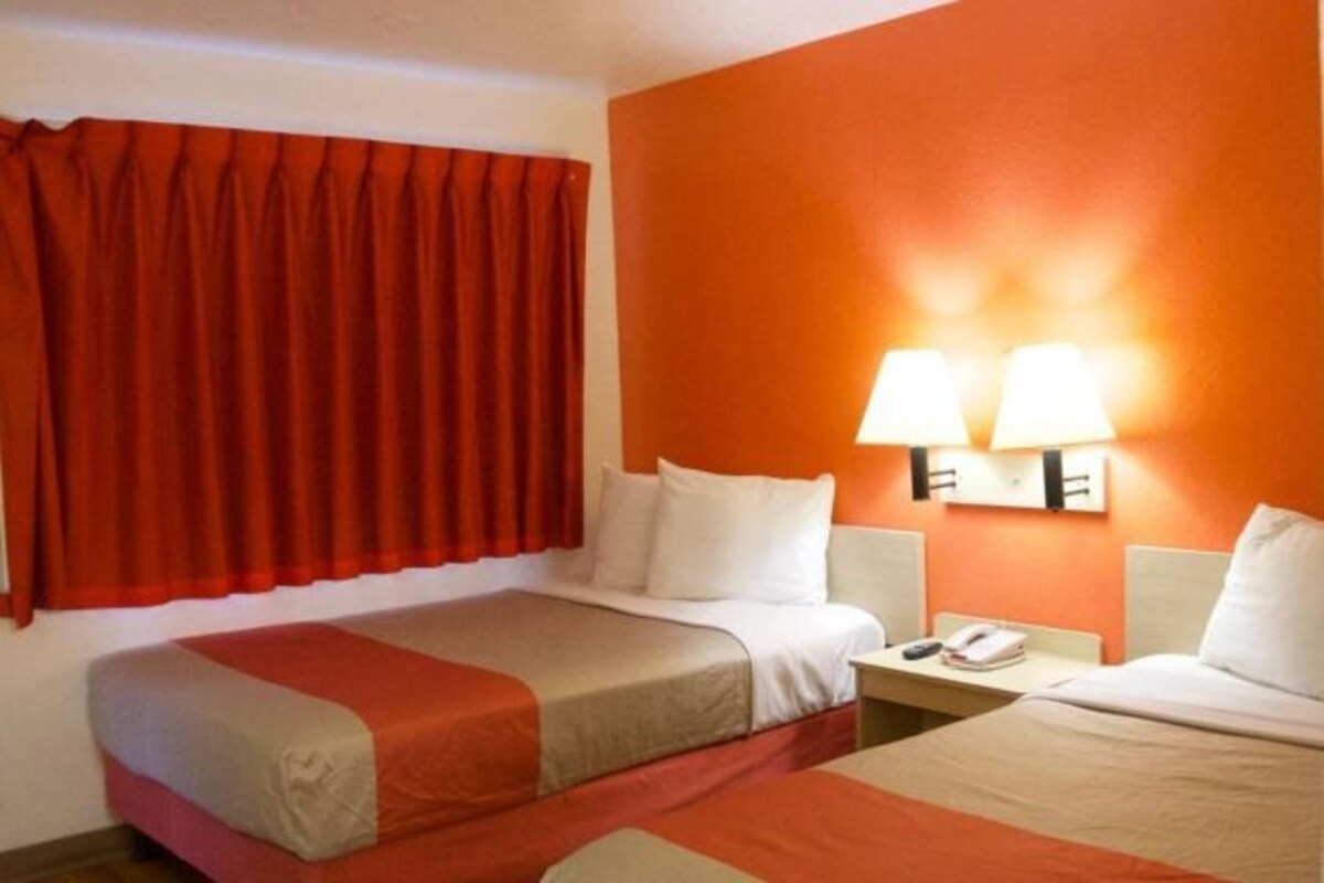 A hotel room with two beds, red curtains, an orange wall, and a nightstand with a phone.