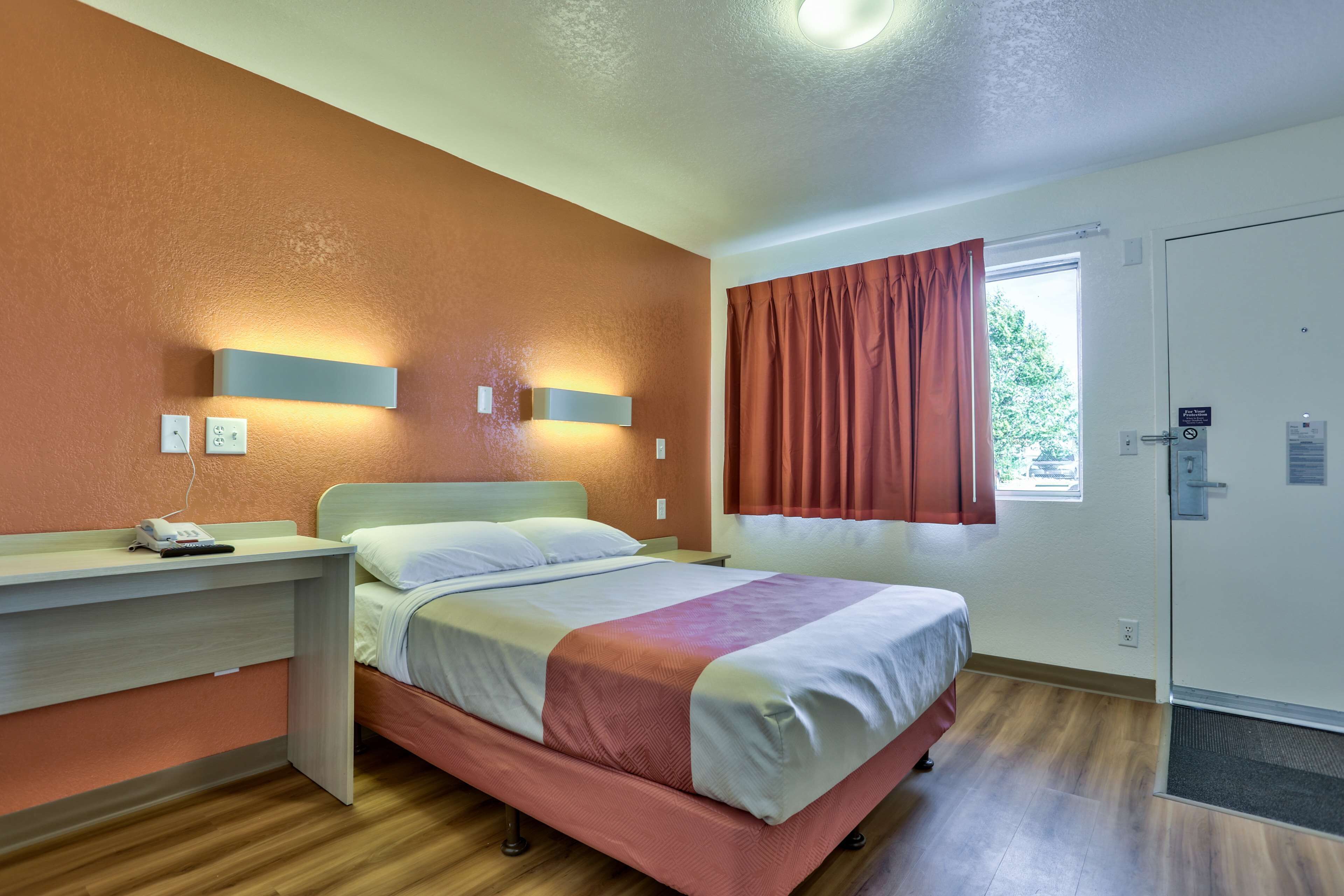 A hotel room with a bed, bedside table, wall-mounted light, window with curtains, and a door.