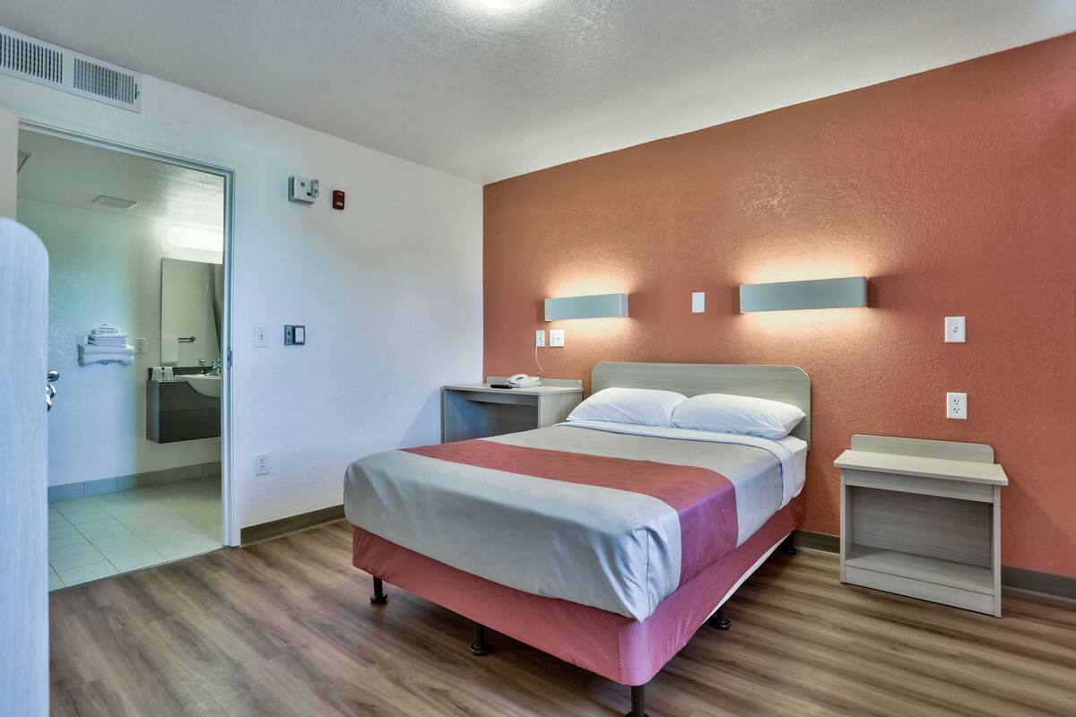 A hotel room with a bed, bedside tables, a mirror, and a bathroom visible through an open door.