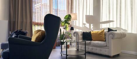 A modern living room with a dark armchair, a white sofa with yellow cushions, a glass coffee table, and a floor lamp.