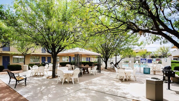An outdoor patio area with tables and chairs, surrounded by trees and a building in the background. - Motel 6 Holbrook, AZ (Holbrook)
