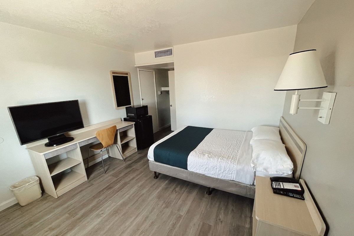 Una habitación de hotel con una cama, un escritorio con televisor, una silla, una lámpara y un pequeño cubo de basura.