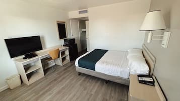 A hotel room with a bed, a desk with a television, a chair, a lamp, and a small trash bin.