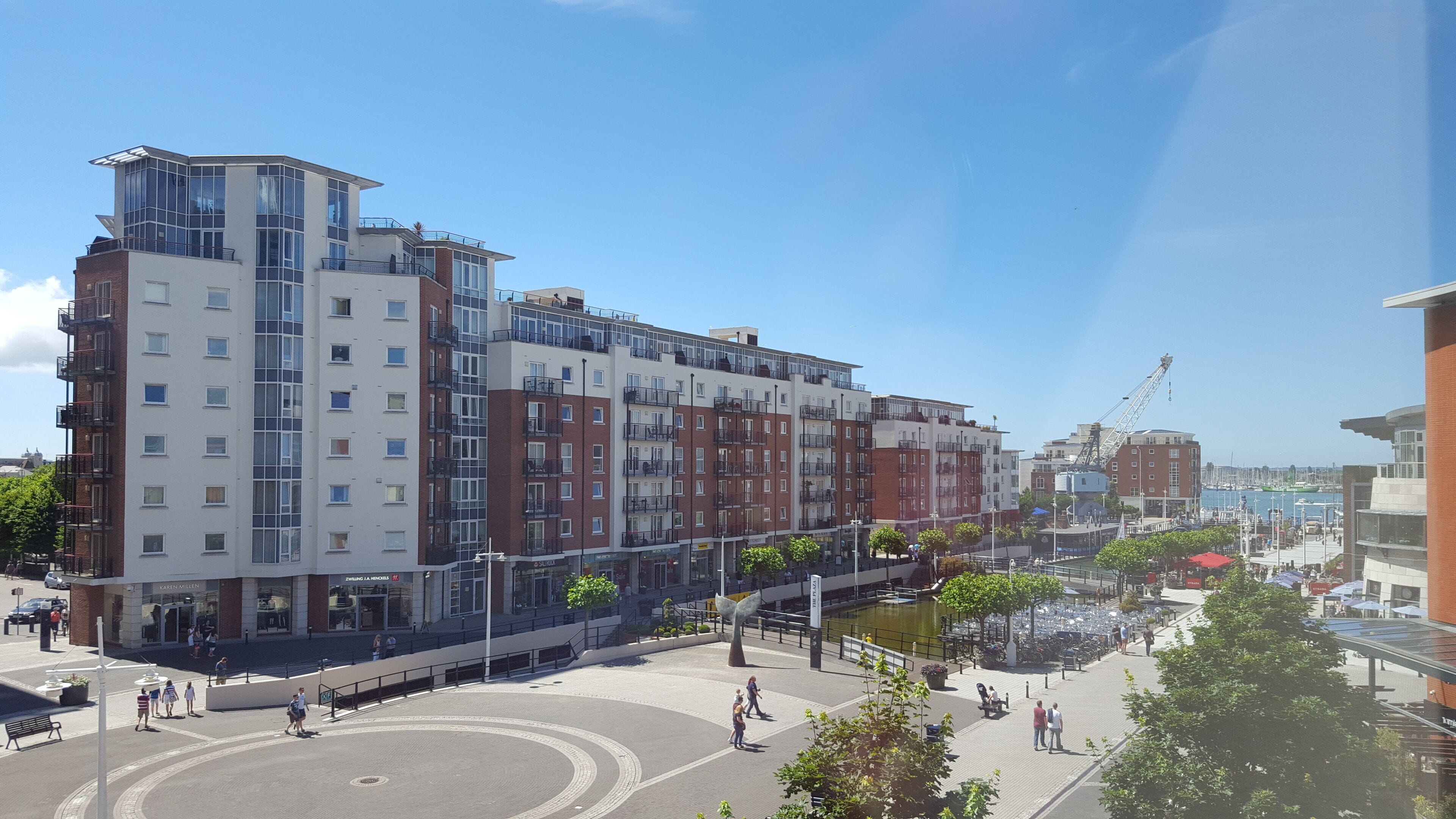 Un immeuble moderne à plusieurs étages, comprenant des logements et des espaces commerciaux, offrant une vue sur le front de mer et doté d’une place publique agréable pour les piétons, avec des espaces aménagés pour s’asseoir.