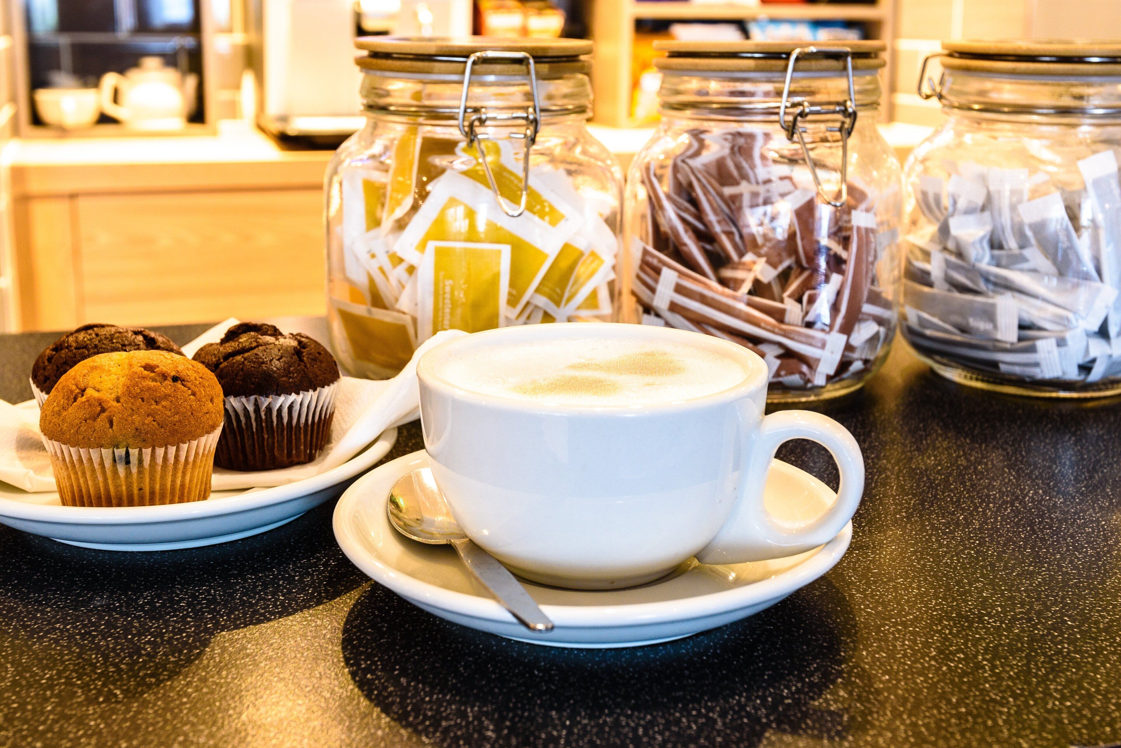 A table with a cup of coffee, a muffin, and jars of tea and chocolate.