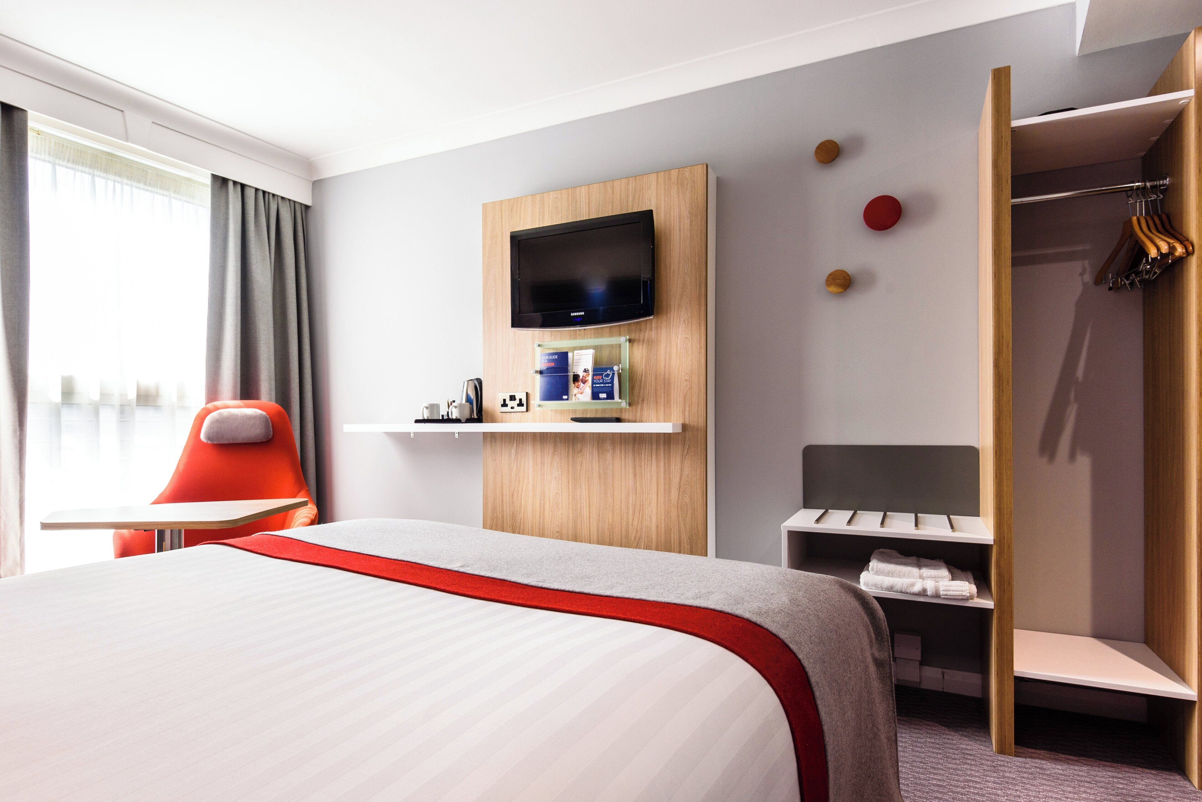 Habitación de hotel con cama, televisor montado en un panel de madera, escritorio con silla, armario y ventana con cortinas.