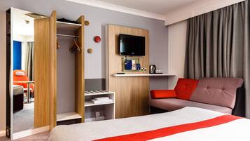 Habitación de hotel con cama, televisor, armario, zona de estar y una mesita con tetera.