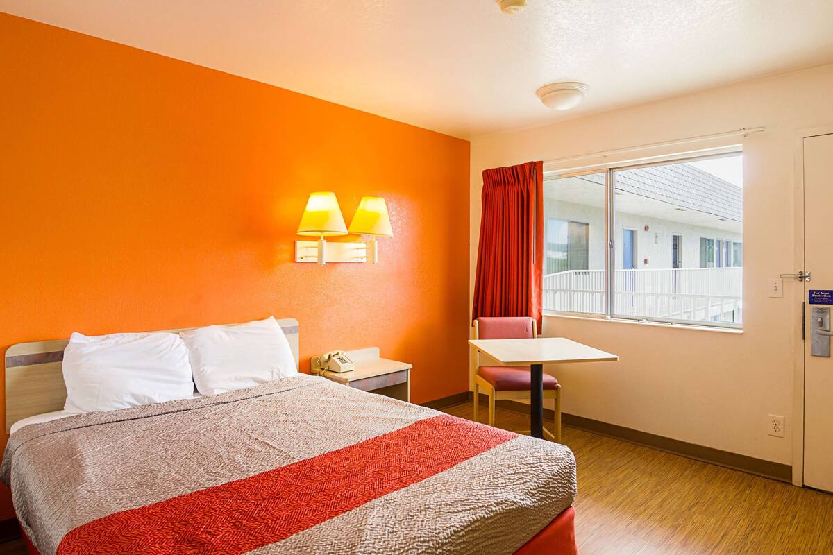 A hotel room with a bed, a desk, a chair, a window with curtains, and a wall-mounted lamp.