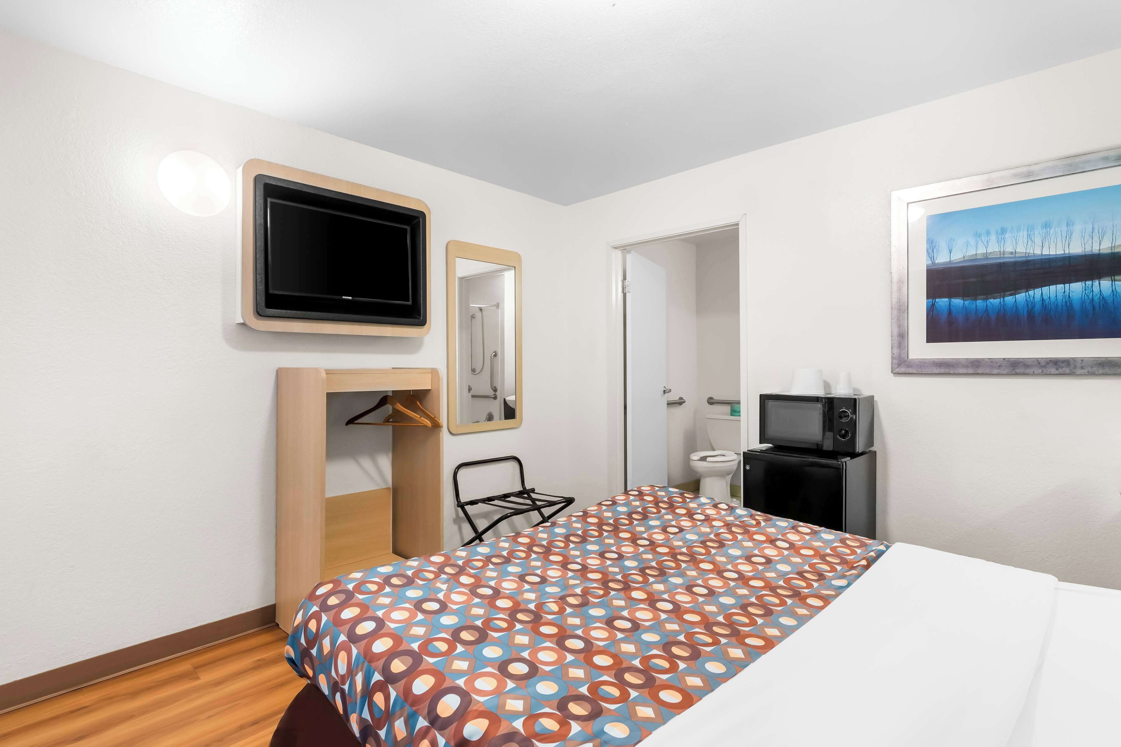 A hotel room with a bed, a television, a mirror, a painting, a wardrobe, and a small desk with a microwave.
