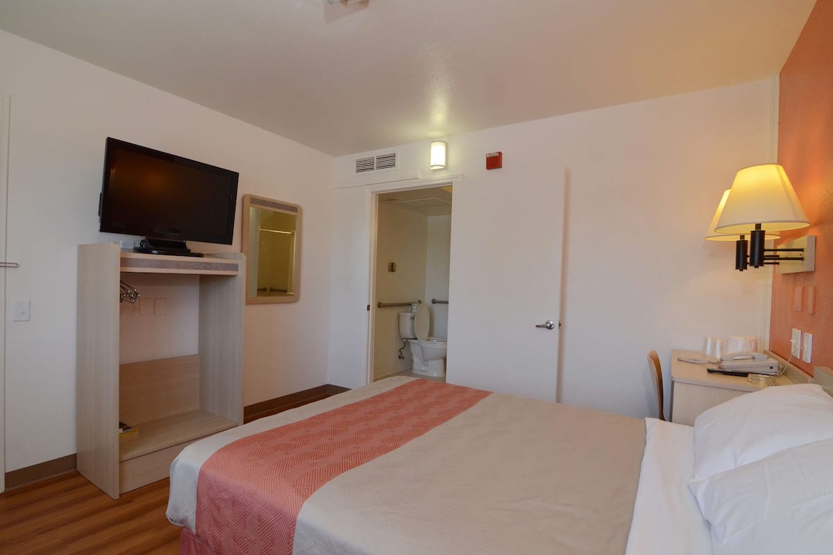 A hotel room with a bed, a TV mounted on a cabinet, a bedside table with a lamp, and a bathroom visible through an open door.