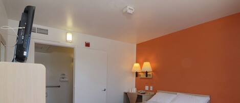 A hotel room with a bed, a television on a stand, a door leading to a bathroom, and two wall-mounted lamps.
