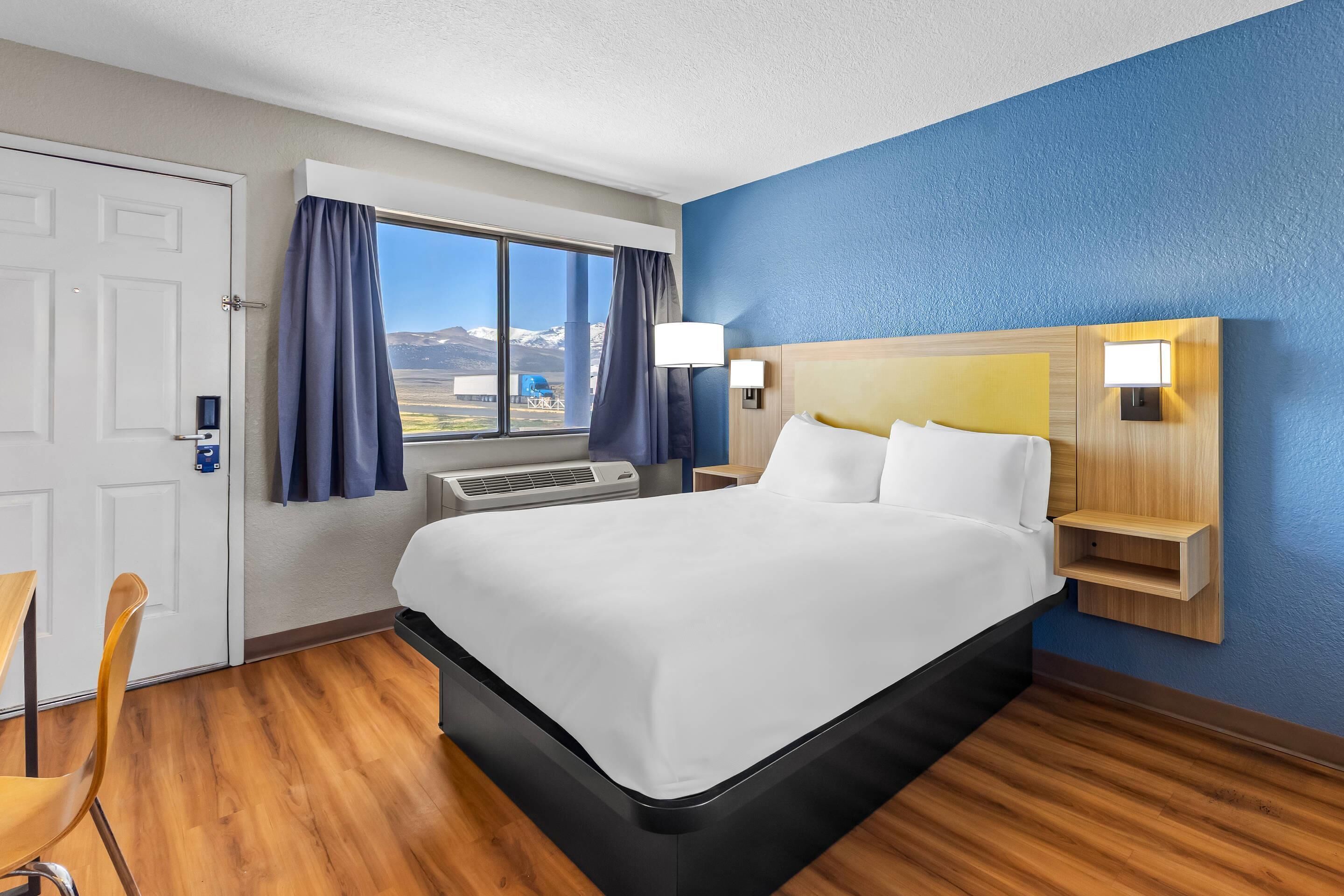 Habitación de hotel con cama, escritorio, silla, ventana con cortinas y vista a las montañas.