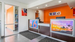 Een hotel receptie met een glazen ingang, een receptiebalie en een muur met ingelijste foto's en een inspirerende quote.