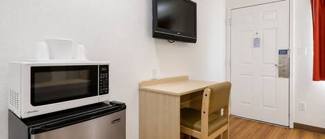 Una kitchenette compacta con microondas, refrigerador y una pequeña mesa con una silla. Un televisor está montado en la pared sobre la mesa.