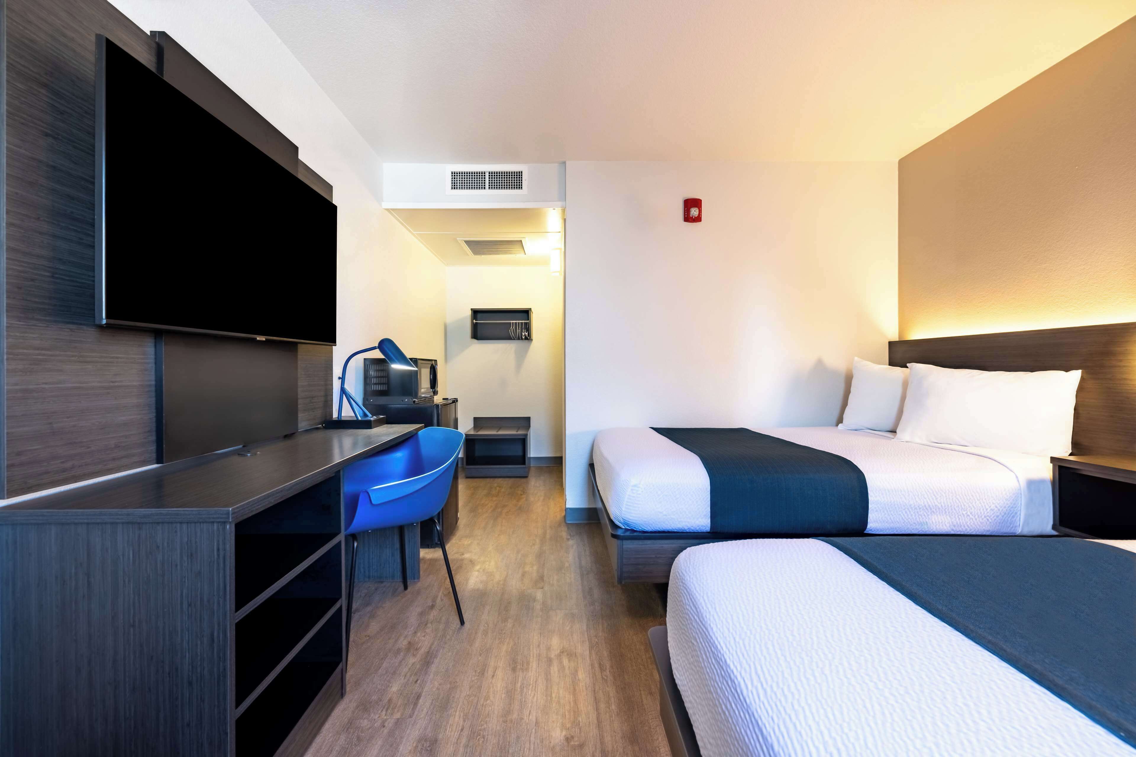 Habitación de hotel con televisor de pantalla plana montado en la pared, un escritorio con silla, dos camas con ropa de cama blanca y oscura, y una pequeña zona de estar con una silla azul.