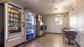 Un interior limpio y moderno con máquinas expendedoras, un dispensador de agua y un pequeño espacio de descanso con mesas y sillas.