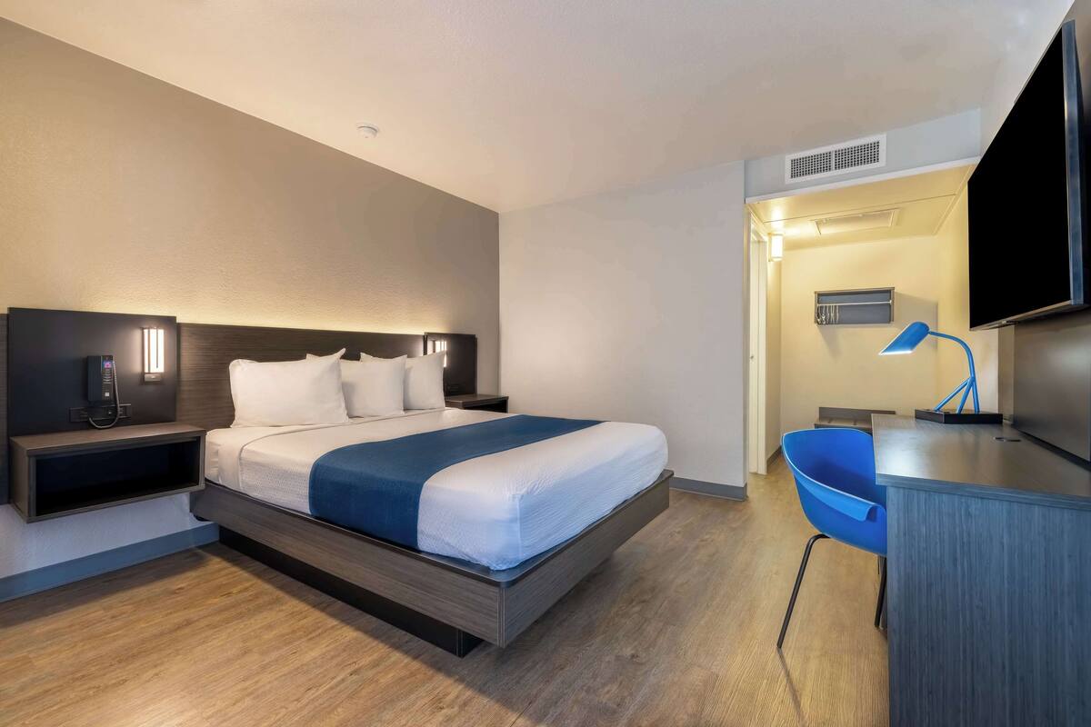 A modern hotel room with a bed, bedside table, lamp, and a desk with a chair.