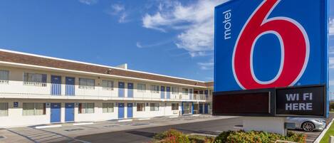 A motel with a large blue sign displaying the number 6, a parking lot, and a sign indicating WiFi availability.