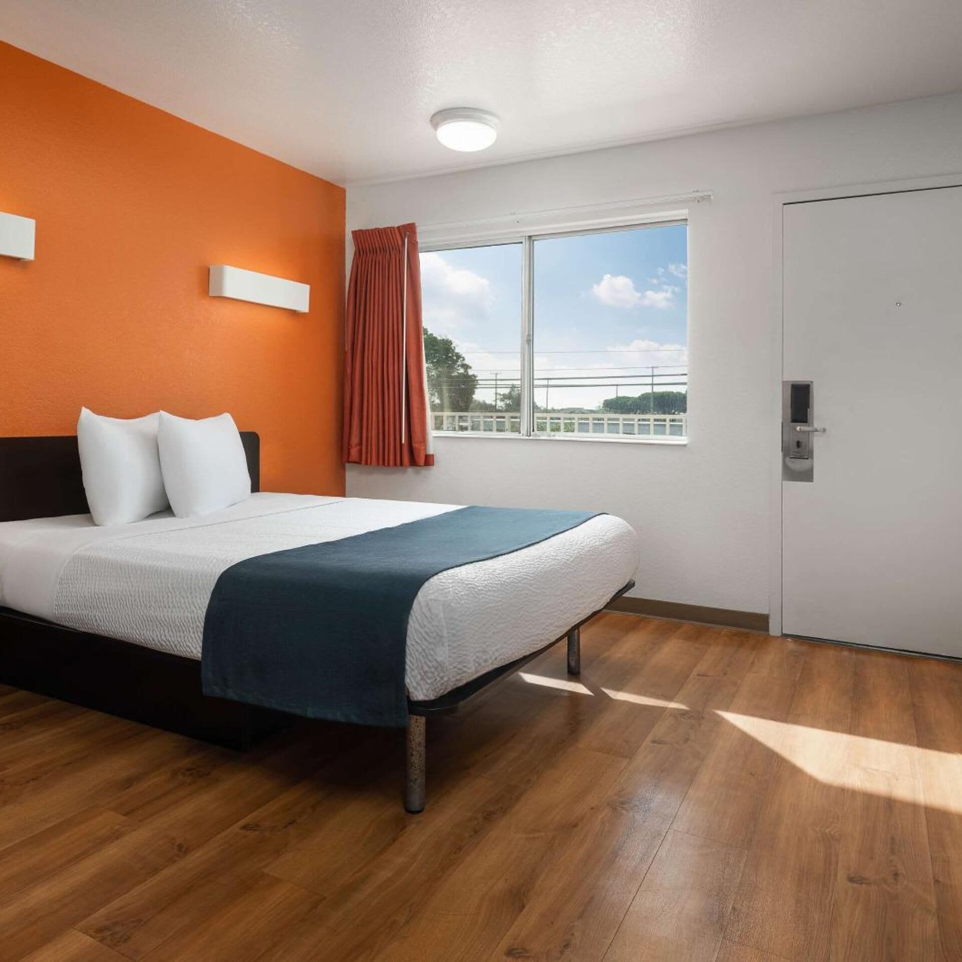 A hotel room with a bed, a window with a view, a door, and a wall-mounted light fixture.