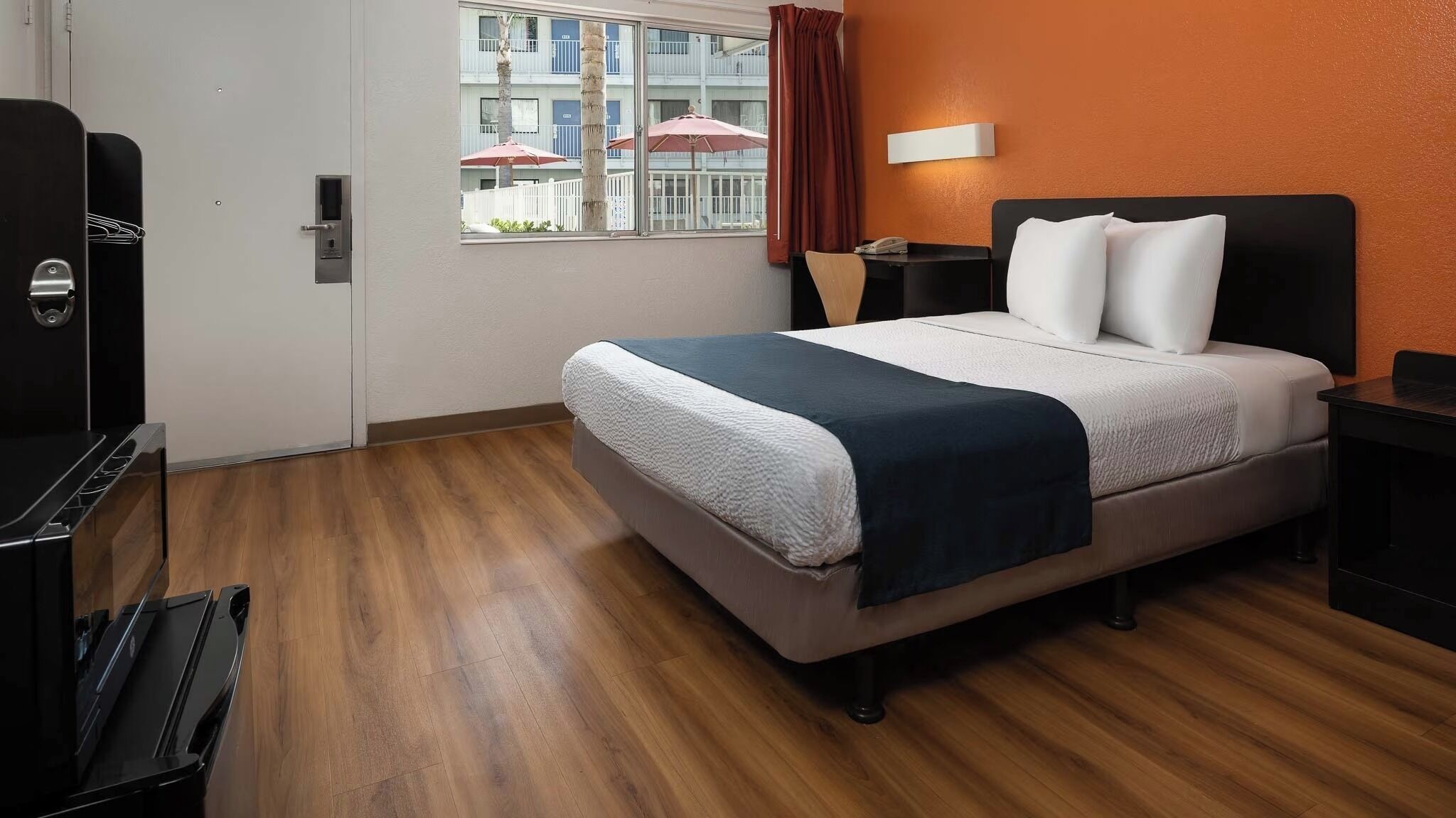 A hotel room with a bed, a desk, a window with curtains, and a view of a building with balconies.