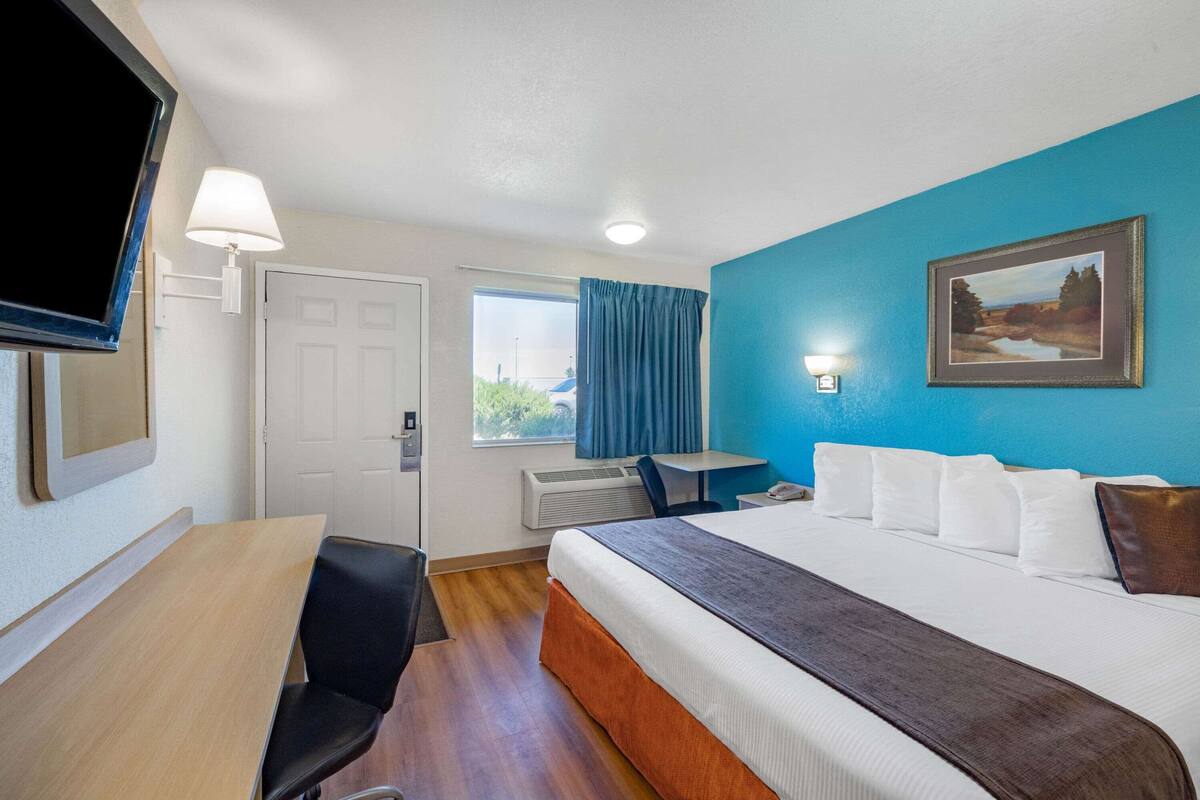 A hotel room with a bed, a desk, a chair, a TV, a window with curtains, and a painting on the wall.