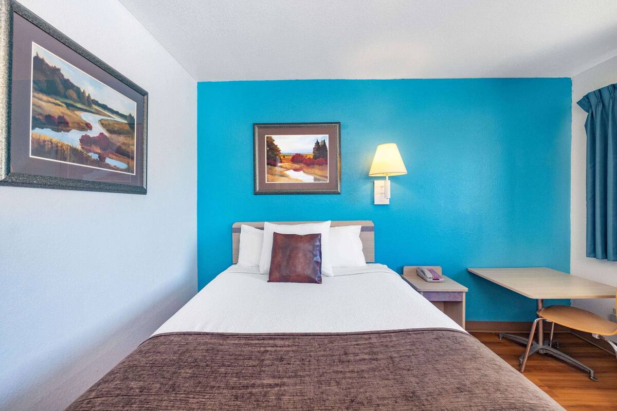 A hotel room with a bed, a desk, a chair, a lamp, and two framed pictures on the wall.