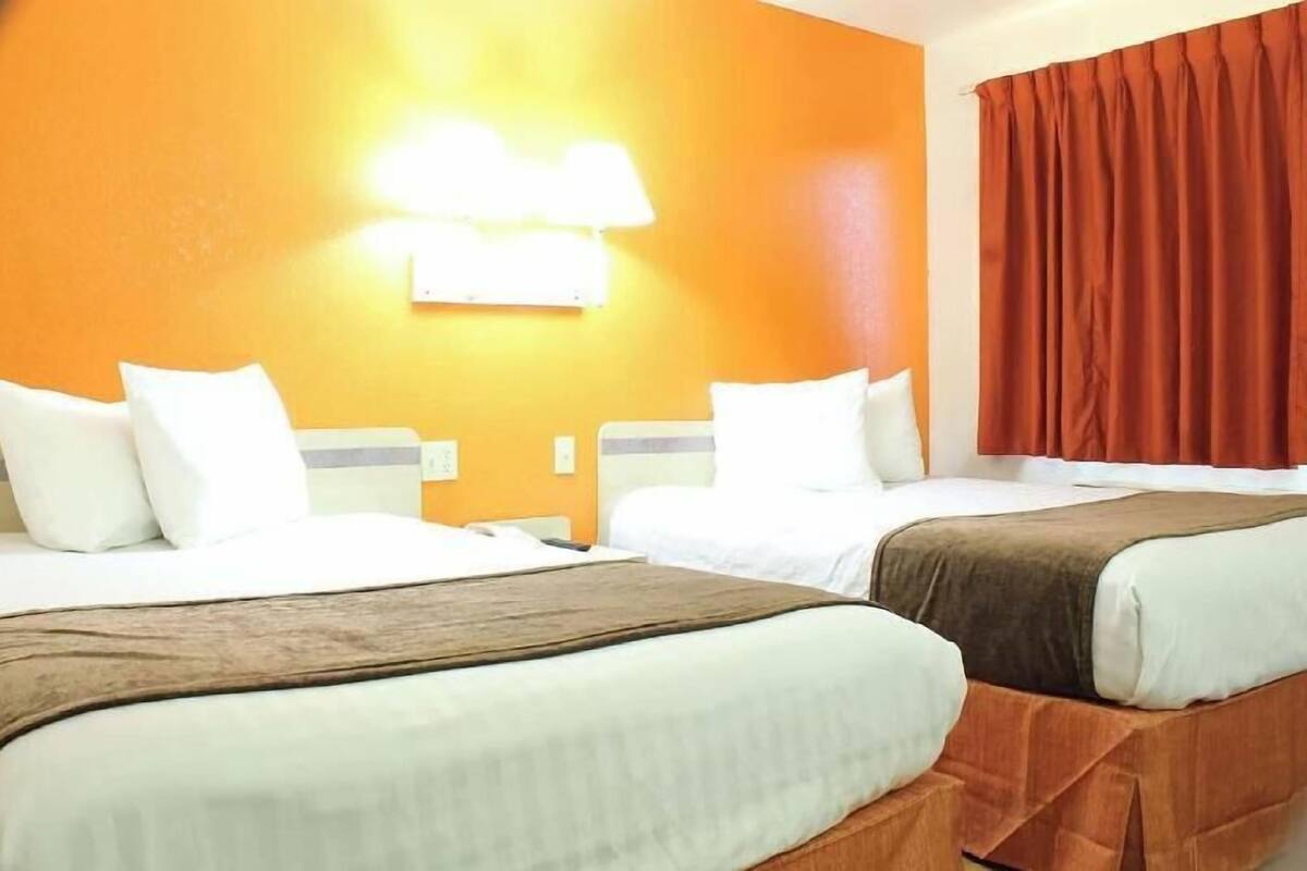 A hotel room with two beds, a bright orange wall, a lamp, and a red curtain.