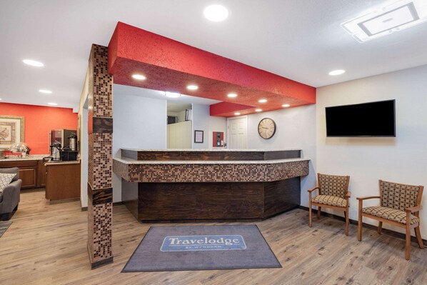 A reception area with a tiled counter, two wooden chairs, a clock, and a flat-screen TV on the wall. - Travelodge by Wyndham Miles City (Miles City)